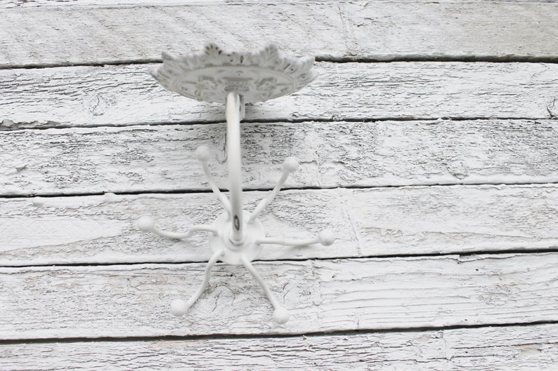 May include: White metal coat rack with a decorative top and five hooks. The rack is mounted on a white wooden wall.