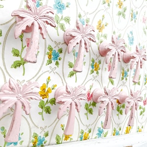 May include: A collection of pink palm tree-shaped cabinet knobs mounted on a floral wallpaper background. The knobs are made of metal and have a distressed finish, adding a vintage touch to the decor.