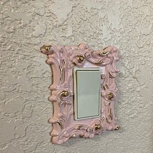 May include: A pink and gold decorative light switch cover with an ornate design. The cover is made of ceramic and has a white light switch plate.