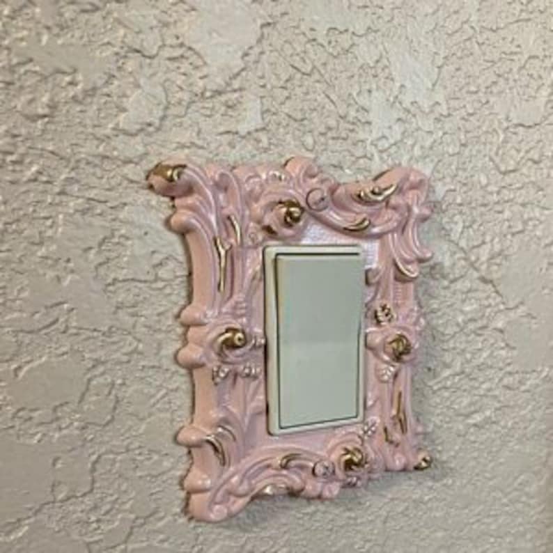 May include: A pink and gold decorative light switch cover with an ornate design. The cover is made of ceramic and has a white light switch plate.