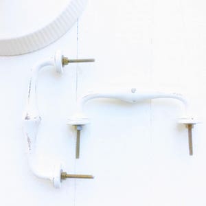 May include: Two white metal door handles with brass screws. The handles are in the shape of a loop with a curved end.