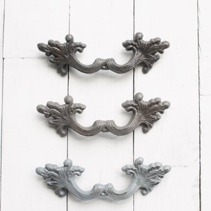 May include: Three ornate metal drawer pulls in different shades of gray. The pulls have a decorative, floral design and are arranged in a vertical row.