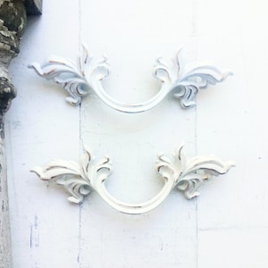May include: Two white ornate drawer pulls with a decorative floral design. The pulls are in the shape of a half-circle with a curved handle.