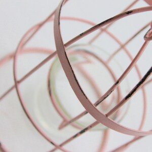 May include: A pink metal birdcage with a distressed finish. The cage has a round base and a cylindrical shape. The cage is empty.