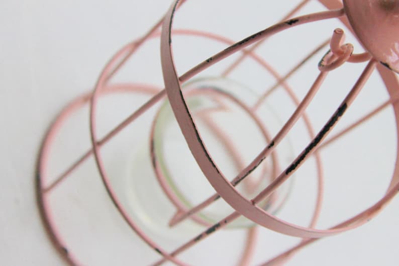 May include: A pink metal birdcage with a distressed finish. The cage has a round base and a cylindrical shape. The cage is empty.