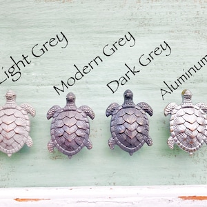 May include: Four decorative turtle-shaped cabinet knobs in varying shades of gray and silver. The knobs are arranged in a row and labeled "Light Grey", "Modern Grey", "Dark Grey", and "Aluminum Silver". The knobs are on a light green wooden surface.