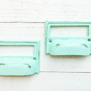 May include: Two light blue metal drawer pulls with a rectangular cutout for a label. The pulls have a slightly distressed finish.