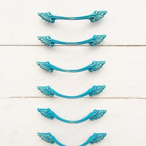 May include: Six turquoise blue drawer pulls with gold accents and ornate floral designs. The pulls are arranged vertically on a white wooden surface.