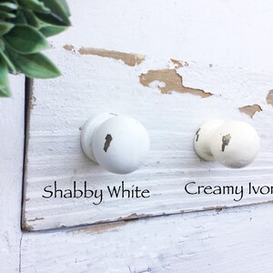May include: Two white knobs with different shades of white. The knob on the left is labeled "Shabby White" and the knob on the right is labeled "Creamy Ivory".