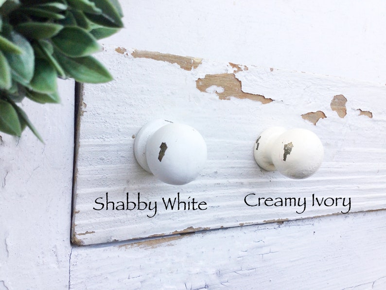 May include: Two white knobs with different shades of white. The knob on the left is labeled "Shabby White" and the knob on the right is labeled "Creamy Ivory".