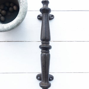 May include: A dark brown, cast iron door pull with an ornate, vintage design. The handle features a long, textured grip with decorative elements at the top and bottom. Two mounting plates with screw holes are visible. The pull is set against a white background.