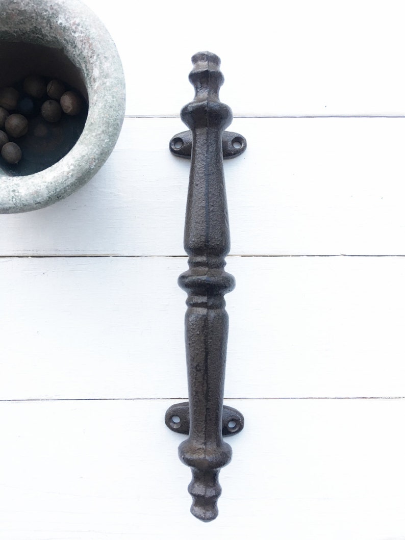 May include: A dark brown, cast iron door pull with an ornate, vintage design. The handle features a long, textured grip with decorative elements at the top and bottom. Two mounting plates with screw holes are visible. The pull is set against a white background.