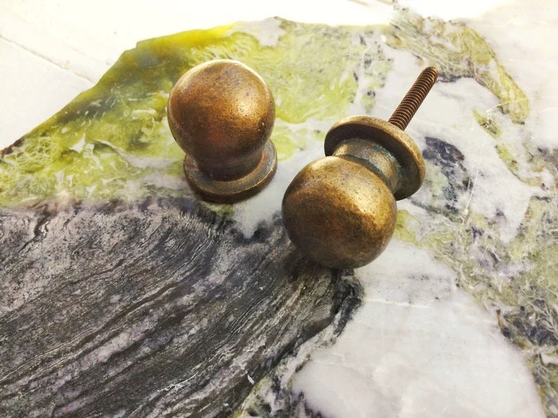 May include: Two antique brass cabinet knobs with a round shape and a slightly raised base. The knobs are on a marble surface.