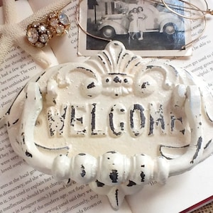 Puede incluir: Un letrero de bienvenida de hierro fundido blanco con un dise&ntilde;o decorativo y un asa. El letrero tiene un acabado desgastado y la palabra "WELCOME" est&aacute; pintada en negro.