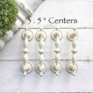 May include: Four white, ornate cabinet pulls with a 3.5 inch center to center spacing. The pulls have a distressed finish and are mounted on a white wooden surface.