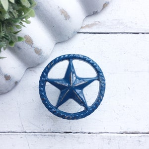 May include: A distressed blue star-shaped cabinet knob with a rope-like border. The knob is centered on a white wooden surface, with a decorative white scalloped edge and greenery in the background.