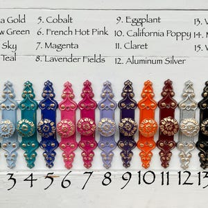 May include: A collection of 15 decorative cabinet pulls in various colors, including yellow, green, blue, pink, and silver. Each pull features a decorative knob and a rectangular backplate. The colors are labeled with numbers and names.
