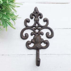 May include: A black cast iron wall hook with a decorative, ornate design. The hook is shaped like a stylized leaf with a single hook at the bottom.