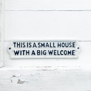 May include: White sign with black lettering that reads "This is a small house with a big welcome". The sign is mounted on a white wooden surface.