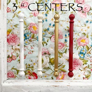 May include: Four decorative cabinet knobs with a 3" center spacing. The knobs are made of metal and have a variety of finishes, including white, gold, and red.