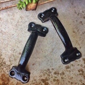 May include: Two black metal door handles with a simple design. The handles have a rounded shape and are attached to a flat surface.