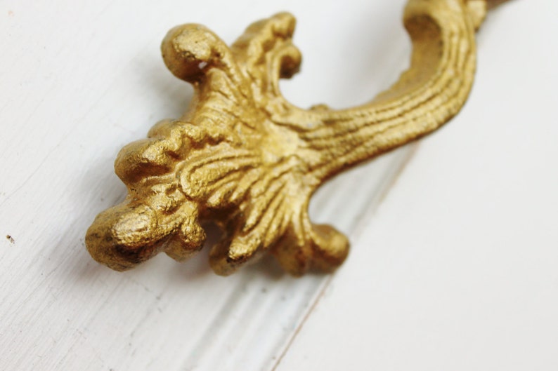 May include: A gold-colored ornate metal drawer pull with a decorative, scroll-like design.