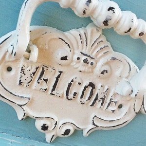 Puede incluir: Un llamador de puerta de metal blanco con un dise&ntilde;o decorativo y la palabra "WELCOME" en relieve en la parte delantera. El llamador tiene un acabado desgastado y est&aacute; fijado a una superficie azul.