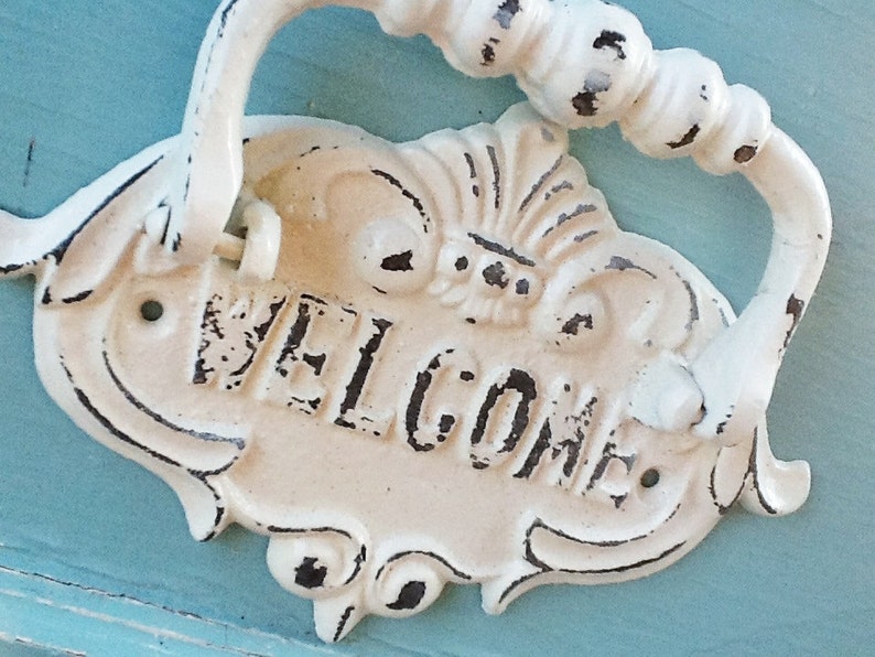 Puede incluir: Un llamador de puerta de metal blanco con un dise&ntilde;o decorativo y la palabra "WELCOME" en relieve en la parte delantera. El llamador tiene un acabado desgastado y est&aacute; fijado a una superficie azul.