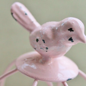May include: A pink bird figurine with black accents sits atop a pink metal birdcage. The birdcage has a round base and multiple metal bars.