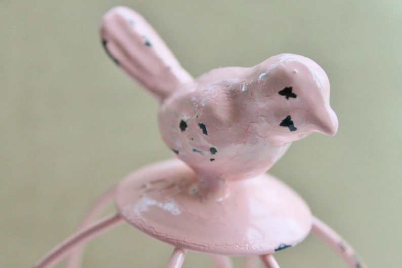 May include: A pink bird figurine with black accents sits atop a pink metal birdcage. The birdcage has a round base and multiple metal bars.