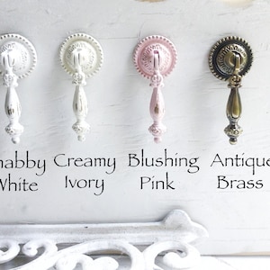 May include: Four decorative cabinet knobs in different finishes: shabby white, creamy ivory, blushing pink, and antique brass. Each knob features a detailed design with a round base and a decorative handle.