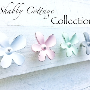 May include: Four flower-shaped metal knobs in white, pink, light green, and gray. The knobs are arranged in a row on a white wooden surface. The text "Shabby Cottage Collection" is written above the knobs.