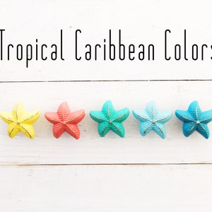 May include: Five starfish in a row on a white wooden surface. The starfish are painted in shades of yellow, orange, turquoise, blue, and light blue. The text "Tropical Caribbean Colors" is written above the starfish.