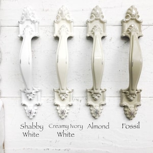 May include: Four ornate cabinet knobs in different colors: shabby white, creamy ivory white, almond, and fossil. The knobs have a decorative, ornate design with a raised, curved handle.