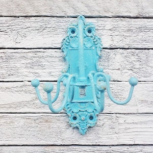 May include: A light blue metal coat rack with ornate details and five hooks. The rack is mounted on a white wooden wall.