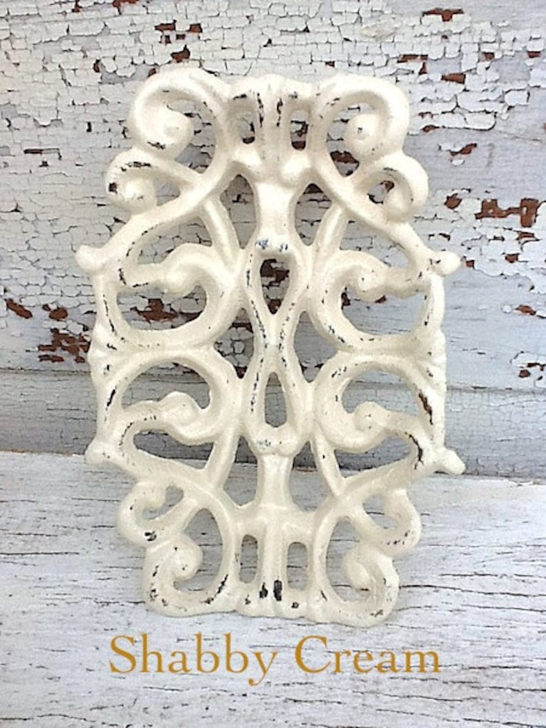 May include: A white, ornate, cast iron decorative piece with a distressed finish. The piece has a delicate, floral design and is reminiscent of a vintage style. The text "Shabby Cream" is visible at the bottom of the image.