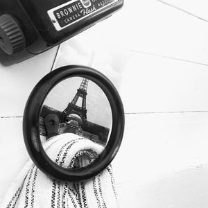May include: A black and white photo of the Eiffel Tower reflected in a round mirror. The mirror is hanging on a white wall. A vintage camera is hanging above the mirror.