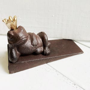 May include: A brown cast iron doorstop shaped like a frog wearing a gold crown. The frog is lying on its side with its legs outstretched.
