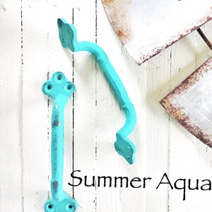 May include: Two turquoise blue metal door handles with decorative details. The handles are on a white wooden background. The text "Summer Aqua" is written below the handles.