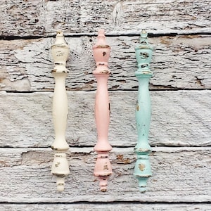 May include: Three decorative cabinet knobs in pastel colors. The knobs are painted in a distressed style with a white, pink, and light blue finish. The knobs have a classic design with a rounded top and a fluted base.