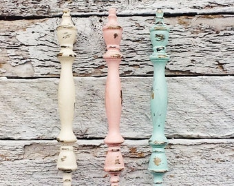 Op de afbeelding: Drie decoratieve kastknoppen in pastelkleuren. De knoppen zijn geschilderd in een verweerde stijl met een witte, roze en lichtblauwe afwerking. De knoppen hebben een klassiek ontwerp met een afgeronde bovenkant en een geribbelde basis.