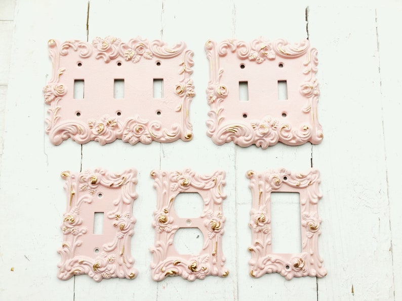 May include: Set of five decorative light switch and outlet covers in a pink and gold colour scheme. The covers feature a detailed ornate design with floral accents.