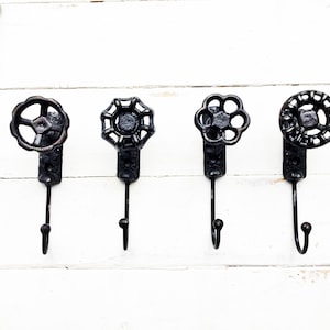 May include: Four black metal hooks with decorative round tops in the shape of vintage water valve handles. The hooks are mounted on a white wooden wall.
