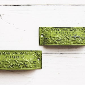 May include: Two green metal drawer pulls with an ornate design. The pulls have a distressed finish and are perfect for adding a vintage touch to any home decor project.