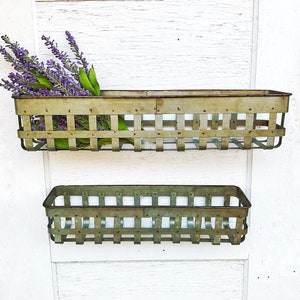 May include: Two metal wall mounted planters with a rustic finish. The top planter is larger and holds a bouquet of purple flowers. The bottom planter is smaller and is empty.