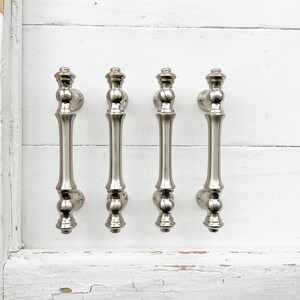 May include: Four silver cabinet door pulls with a decorative design. The pulls are mounted on a white painted wall.