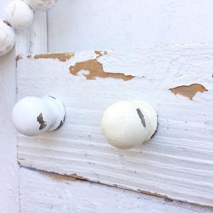 May include: Two white knobs with a distressed finish are attached to a white painted wooden surface. The paint is chipped and peeling, revealing the natural wood underneath.