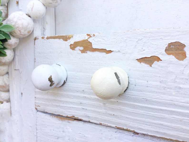 May include: Two white knobs with a distressed finish are attached to a white painted wooden surface. The paint is chipped and peeling, revealing the natural wood underneath.
