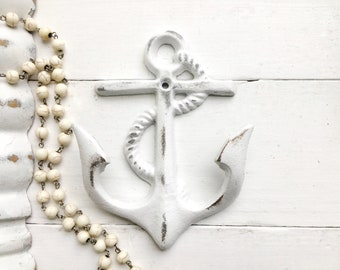 May include: White painted metal anchor with a rope design. The anchor is on a white wooden background.