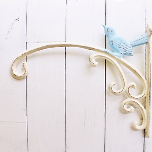 May include: White metal wall-mounted plant hanger with a decorative scroll design and a light blue bird figurine.